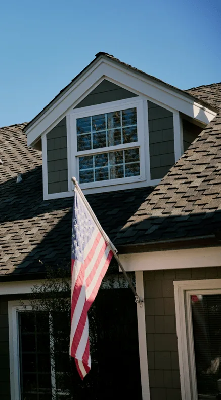 American flag mounted on the exterior of a gray house with a dormer window and shingled roof