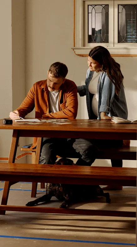 Pregnant woman and man reviewing documents at a wooden dining table with a dog resting underneath