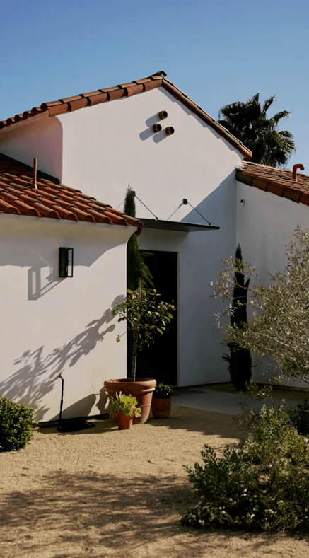 Spanish Colonial-style white stucco home with terra cotta tile roof, black front door, and potted plants in a sunny courtyard