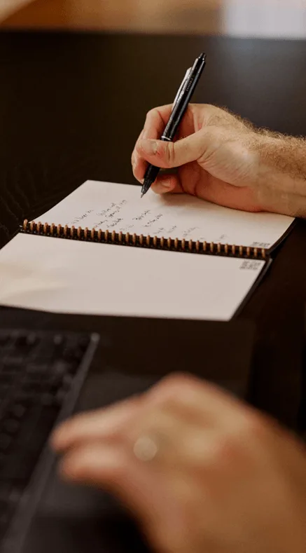 Person writing notes in a spiral notebook with a pen, with a laptop keyboard visible in the foreground