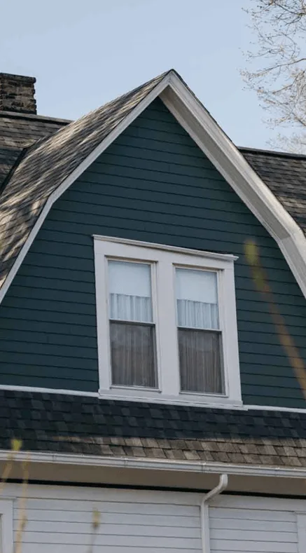 Upper story of a house with dark green siding, white-trimmed double window, and a gambrel roof with aged shingles
