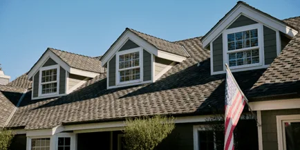 American flag mounted on the exterior of a gray house with a dormer window and shingled roof