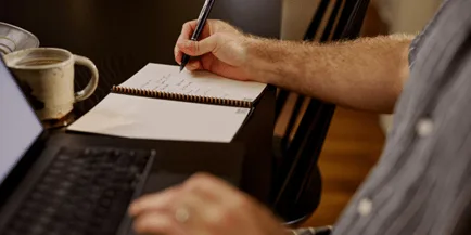 Person writing notes in a spiral notebook with a pen, with a laptop keyboard visible in the foreground