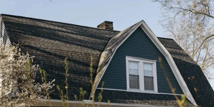 Upper story of a house with dark green siding, white-trimmed double window, and a gambrel roof with aged shingles