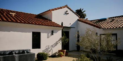 Spanish Colonial-style white stucco home with terra cotta tile roof, black front door, and potted plants in a sunny courtyard